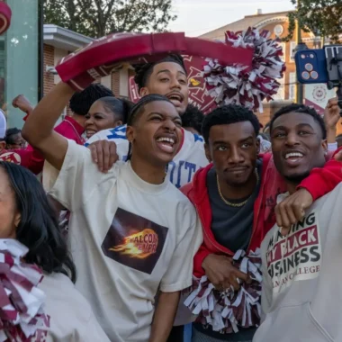 "College students at a lively outdoor event, smiling and cheering with pom-poms and team gear, taking selfies and enjoying a festive, energetic atmosphere on campus."